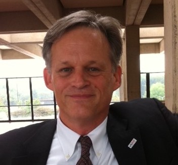 Photograph of white man with gray hair parted to the side wearing black blazer, white collared shirt and dark tie standing outside under a roof